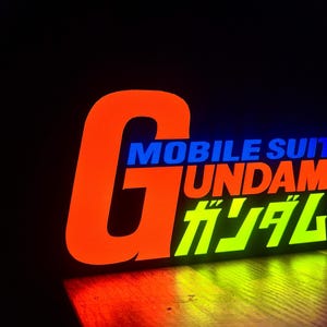 以下が含まれることがあります： オレンジ、青、緑で「MOBILE SUIT GUNDAM」と書かれた光る看板。大きなオレンジ色の「G」が左側にあり、その下に日本語の文字があります。暗い背景の中で看板が光っています。