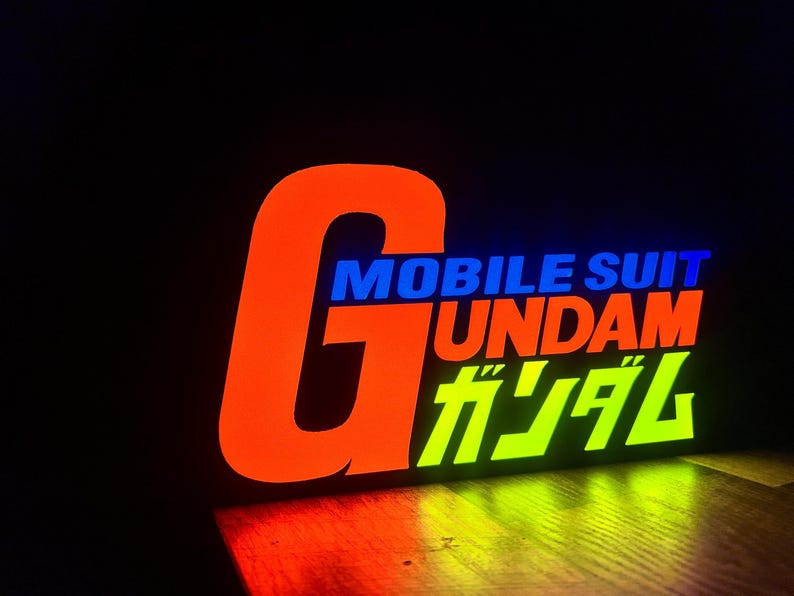 以下が含まれることがあります： オレンジ、青、緑で「MOBILE SUIT GUNDAM」と書かれた光る看板。大きなオレンジ色の「G」が左側にあり、その下に日本語の文字があります。暗い背景の中で看板が光っています。