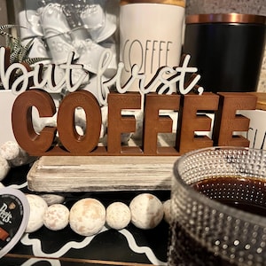 May include: A wooden sign with the phrase "but first, COFFEE" in brown and white lettering. The sign sits on a white and gray base, with a glass of coffee in the foreground.