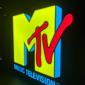 May include: Illuminated MTV logo with a yellow background, red 'MTV' lettering, and blue outlines. The words 'MUSIC TELEVISION' are below the logo in blue. The image is set against a black background.