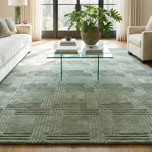 Könnte beinhalten: Ein großer, rechteckiger, salbeigrüner Teppich mit einem geometrischen Muster aus sich kreuzenden Linien. Der Teppich befindet sich in einem Wohnzimmer mit einem cremefarbenen Sofa, Sesseln und einem Glastisch. Der Raum hat große Fenster und eine neutrale Farbpalette.