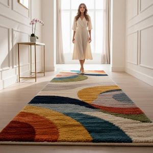 Op de afbeelding: Een kleurrijk, rechthoekig vloerkleed met een abstract ontwerp. Het vloerkleed heeft een patroon van overlappende bogen in tinten oranje, geel, blauw, groen en grijs. Het vloerkleed ligt in een gang met een vrouw die naar de camera loopt.