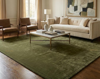 May include: A large, olive green area rug with a subtle textured pattern dominates a living room. The rug is framed by a dark border and complements the room's neutral color palette, including a cream-colored sofa, brown chairs, and a marble coffee table.