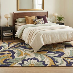 May include: A bedroom scene featuring a bed with a wooden headboard, pillows, and a cream-colored blanket. A patterned rug with blue, yellow, and green floral designs is on the floor. Two nightstands with lamps and books flank the bed.
