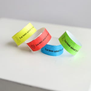 May include: Four colorful paper wristbands in yellow, red, blue, and green, each with the text "I've been asked to..." printed in black. The wristbands are arranged on a white surface.