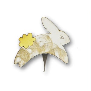 May include: A white bunny-shaped bottle stopper with a yellow flower accent. The bunny has gold swirl details and a silver stem. A decorative item for wine bottles.