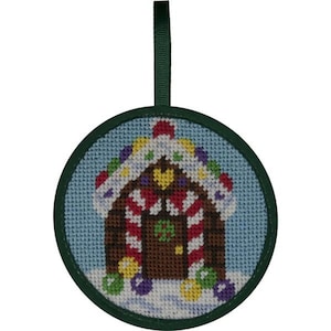May include: Round Christmas ornament featuring a cross-stitched gingerbread house with candy cane trim, colorful lights, and a green door. The ornament has a green border and a green ribbon for hanging. The background is light blue.