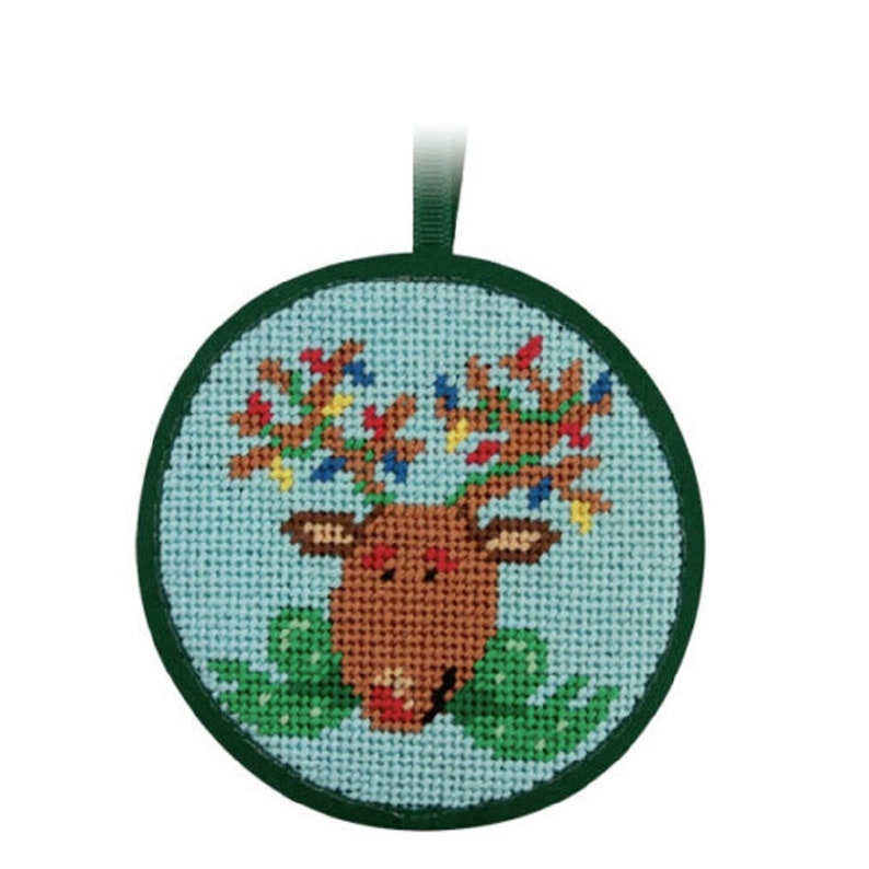 May include: A round Christmas ornament featuring a cross-stitched reindeer design. The reindeer has brown antlers adorned with colorful lights, a brown face, and a green bow. The ornament has a green border and a green ribbon for hanging.
