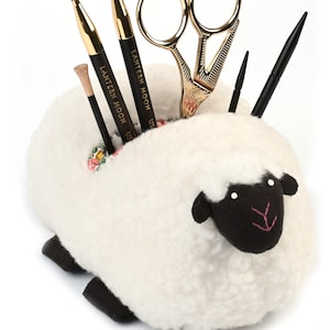 May include: White sheep-shaped needle minder with black eyes and nose, holding a pair of gold scissors, three gold knitting needles, and two black crochet hooks.
