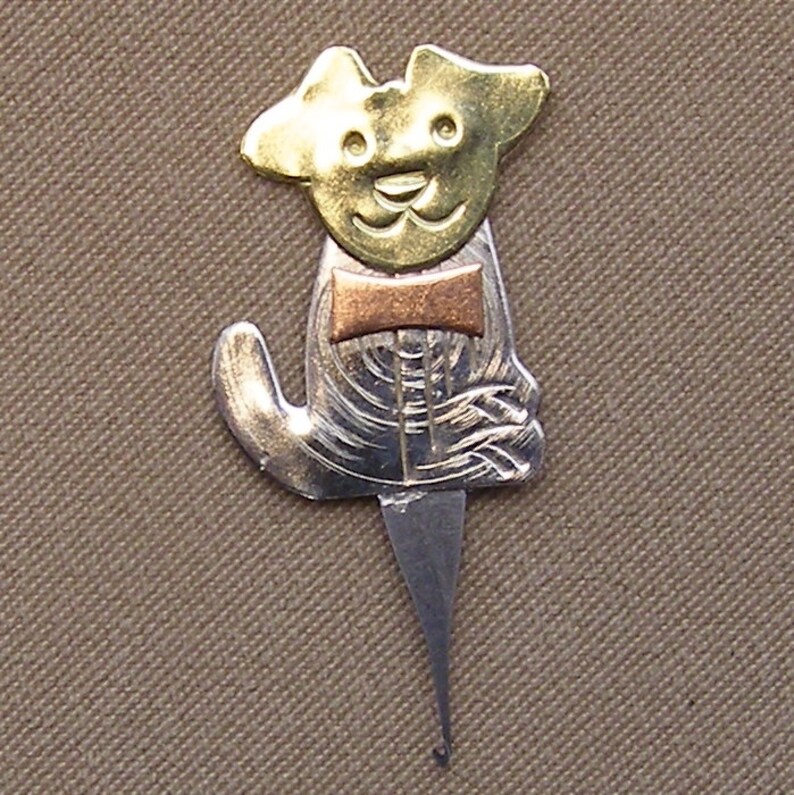 May include: A metal dog-shaped toothpick holder with a gold-colored head, a copper-colored bow tie, and a silver-colored body. The toothpick holder has a pointed end for inserting into a toothpick.