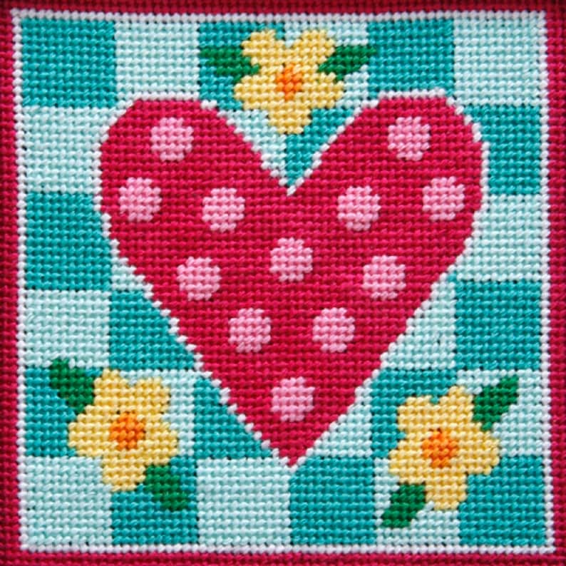Pode incluir: Um cora&ccedil;&atilde;o rosa com bolinhas brancas &eacute; cercado por tr&ecirc;s flores amarelas com folhas verdes em um fundo xadrez azul e branco.
