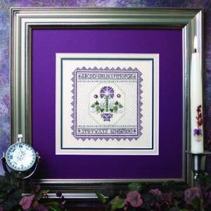 May include: A framed cross-stitch design featuring a purple flower arrangement in a white pot. The design is surrounded by a border with the alphabet and numbers. The frame is silver with a purple mat.