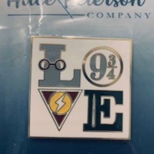 May include: A gold-toned enamel pin featuring a stylized "LOVE" design. The "L" is a pair of glasses, the "O" is a circle with the number "9 3/4" inside, the "V" is a lightning bolt in a triangle, and the "E" is a vertical line with a needle.