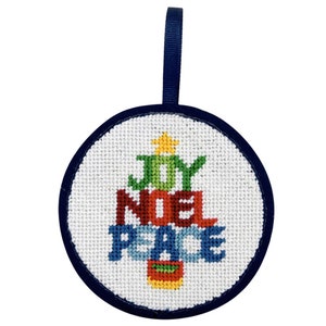 May include: Round Christmas ornament with a white background and a navy blue border. The ornament features a stitched Christmas tree design with the words "JOY NOEL PEACE" in green, red, and blue. A navy blue ribbon is attached for hanging.
