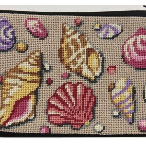 Op de afbeelding: Een beige etui met ritssluiting en zwarte rand, met een kruissteekontwerp van verschillende schelpen in tinten geel, roze, paars en bruin. De schelpen zijn verspreid over het oppervlak.