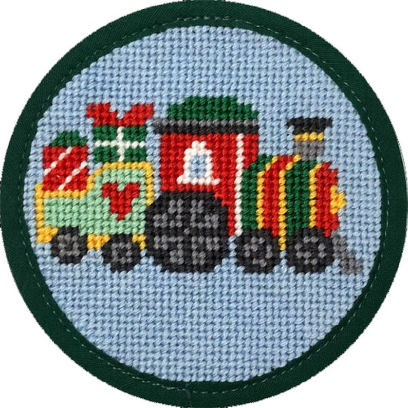 May include: A round, cross-stitched Christmas ornament featuring a colorful train carrying gifts. The train is red, green, yellow, and black, set against a light blue background. The ornament is framed by a dark green border.