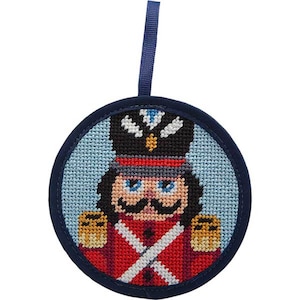 May include: A round, embroidered Christmas ornament featuring a nutcracker soldier. The ornament has a navy blue border and a light blue background. The nutcracker is dressed in a red uniform with gold accents, a black hat, and a large mustache. A navy blue ribbon is attached for hanging.
