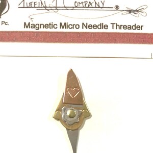 May include: A magnetic micro needle threader with a gnome-like design. The top portion is copper-colored with a heart-shaped cutout. The lower section is silver-colored with a curved hook. The text "Magnetic Micro Needle Threader" is visible.