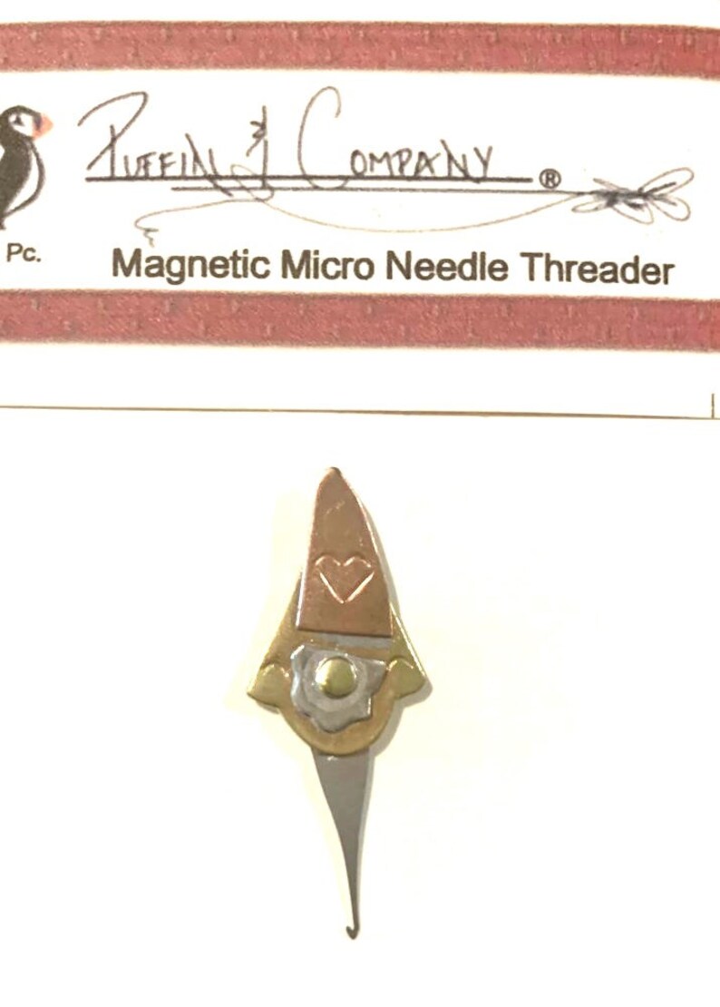 May include: A magnetic micro needle threader with a gnome-like design. The top portion is copper-colored with a heart-shaped cutout. The lower section is silver-colored with a curved hook. The text "Magnetic Micro Needle Threader" is visible.
