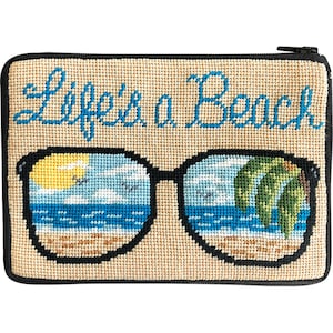 Op de afbeelding: Een beige etui met ritssluiting en naaldwerk met de tekst "Life's a Beach" in blauw script. Het ontwerp toont een zwarte zonnebril met een strandsc&egrave;ne die in de glazen wordt weerspiegeld. De strandsc&egrave;ne omvat een gele zon, blauw water en palmbomen.