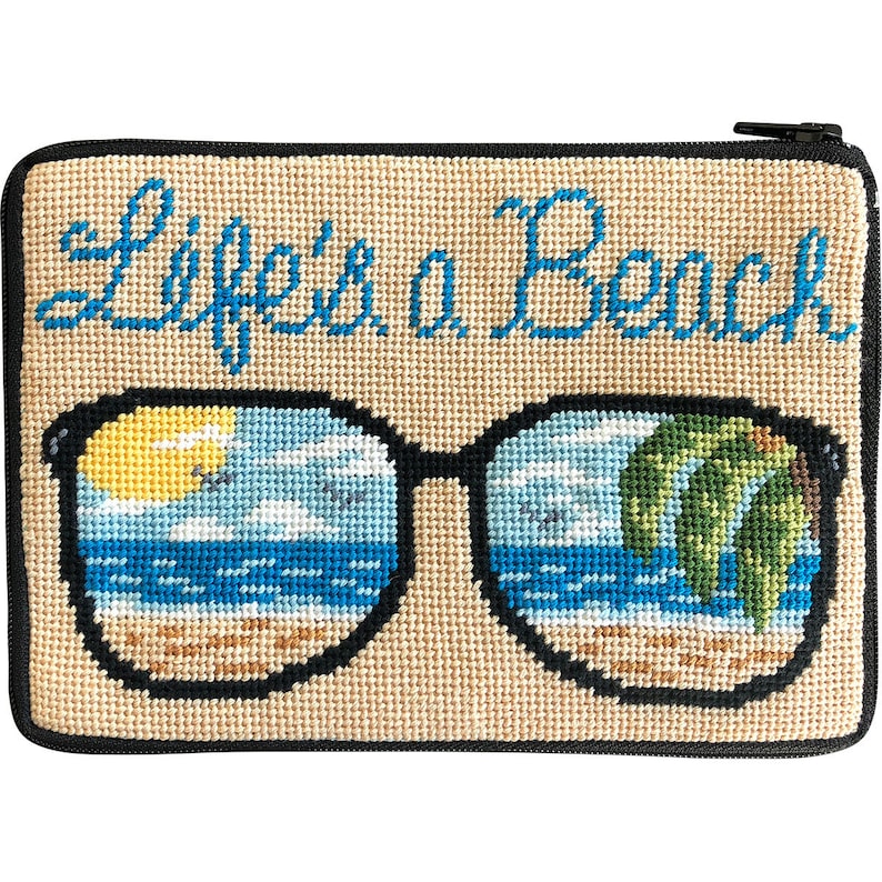Op de afbeelding: Een beige etui met ritssluiting en naaldwerk met de tekst "Life's a Beach" in blauw script. Het ontwerp toont een zwarte zonnebril met een strandsc&egrave;ne die in de glazen wordt weerspiegeld. De strandsc&egrave;ne omvat een gele zon, blauw water en palmbomen.