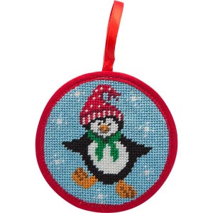 May include: A round, red-trimmed ornament with a blue background and a white penguin wearing a red and white hat and a green scarf. The penguin is standing on a snowy background.