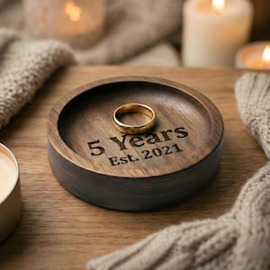 Op de afbeelding: Een ronde, houten ringschaal met een gouden ring erin. De schaal is gegraveerd met "5 Years Est. 2021". De schaal is donkerbruin met een lichtere bruine binnenkant. De achtergrond bevat kaarsen en een gebreide deken.