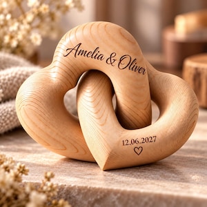 May include: Two interlocking light-colored wooden hearts with a smooth grain finish. The top heart is engraved with the text Amelia & Oliver in a dark script font. The bottom heart features the engraved date 12.06.2027 and a small heart icon.