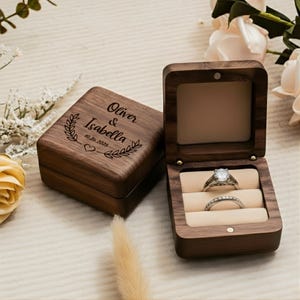 May include: A wooden ring box with the names "Oliver & Isabella" and the date "10.26.2025" engraved on the lid. The open box displays two rings on a beige interior. The box is made of dark brown wood.
