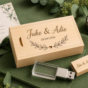 May include: A rectangular light-colored wooden box with a sliding lid engraved with the names Jake & Adie, the date 28/08/2026, and a leaf motif. Beside it lies a clear crystal USB flash drive with a matching wooden cap engraved with the same names.
