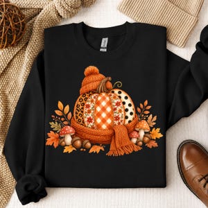 Peut inclure: Un sweat-shirt noir à manches longues avec un motif central représentant une citrouille portant un bonnet et une écharpe en tricot orange. La citrouille présente des sections à motifs léopard, vichy, floraux et à pois, entourée de feuilles d'automne, de champignons et de glands.