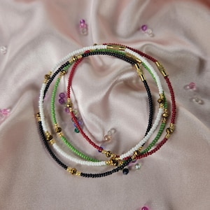 Puede incluir: Una colección de collares de cuentas en varios colores, incluyendo negro, blanco, rojo y verde, con detalles dorados. Los collares están dispuestos sobre una superficie de satén rosa, con cuentas dispersas para un efecto decorativo.