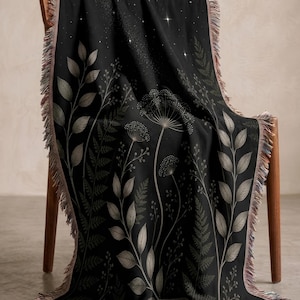 May include: A black woven throw blanket featuring a botanical pattern of light gray ferns, leaves, and dandelion-like seed heads. The top section displays a starry night sky design with small white sparkles. The edges have a multicolored fringe.