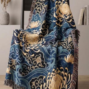 May include: A woven throw blanket featuring a dark blue and black background with a repeating pattern of large gold chrysanthemum flowers and swirling blue wave motifs. The blanket has a fringed edge along the sides and bottom.