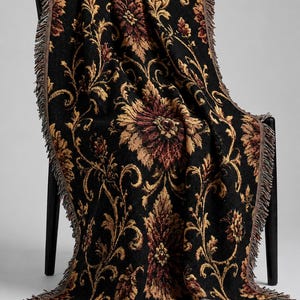 May include: A woven throw blanket featuring a black background with a repeating floral pattern in shades of gold, rust, and deep red. The rectangular textile has decorative fringe along the edges and is draped over a black chair.