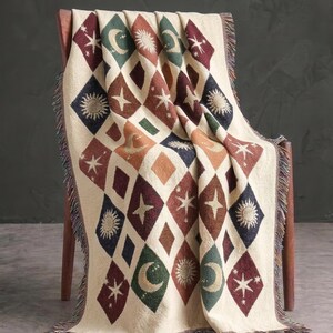 May include: A woven throw blanket featuring a diamond pattern in cream, dark green, deep red, and navy blue. Each diamond contains a celestial motif, including suns, moons, and stars. The blanket has a fringed edge along the sides and bottom.
