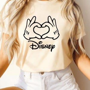 May include: Cream-colored t-shirt featuring a black graphic of Mickey Mouse hands forming a heart shape, with the word "Disney" below. The shirt is made of a soft material.