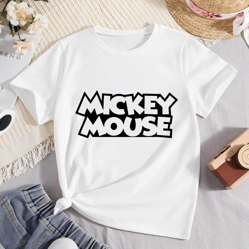 May include: White t-shirt featuring the text "MICKEY MOUSE" in a bold, black, outlined font. The shirt is made of a soft material and has short sleeves. The design is centered on the front of the shirt.