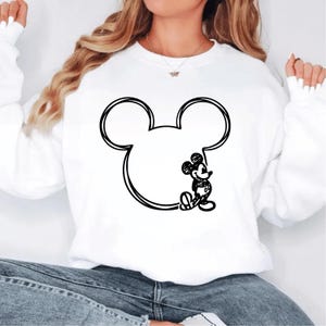 May include: White sweatshirt featuring a large black outline of Mickey Mouse's head and ears. A small, detailed sketch of Mickey Mouse is drawn inside the outline. The sweatshirt is a casual style, suitable for everyday wear.
