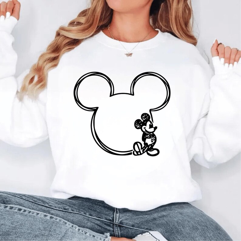 May include: White sweatshirt featuring a large black outline of Mickey Mouse's head and ears. A small, detailed sketch of Mickey Mouse is drawn inside the outline. The sweatshirt is a casual style, suitable for everyday wear.