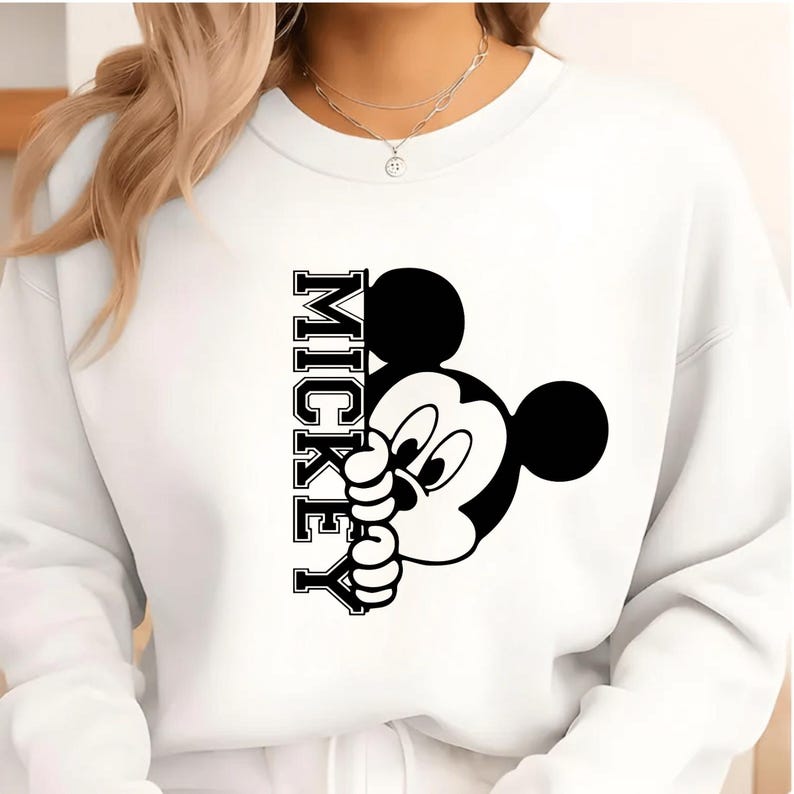 May include: White sweatshirt featuring a black and white graphic of Mickey Mouse peeking out from behind the word "MICKEY" in a vertical, collegiate-style font. The design is centered on the front of the sweatshirt.