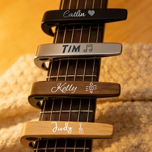 Op de afbeelding: Vier gitaarcapo's in zwart, zilver en houttinten worden weergegeven op de toets van een gitaar. Elke capo is gegraveerd met een naam en een muzieksymbool of hart. De namen zijn "Catlin", "Tim", "Kelly" en "Judy".