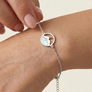 Puede incluir: Una pulsera de plata con un colgante circular que representa una montaña y un sol. El colgante tiene una incrustación de nácar y un pequeño acento dorado. La pulsera está siendo sostenida por una persona.
