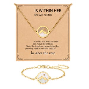 Puede incluir: Un conjunto de collar y pulsera de color dorado. Cada pieza presenta un colgante circular con un diseño de montaña y un fondo blanco. El collar se presenta con una tarjeta que dice "IS WITHIN HER she will not fall" y otro texto inspirador.