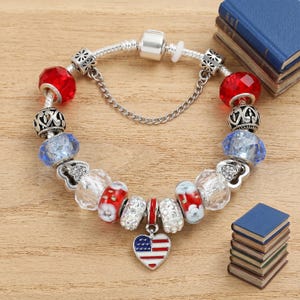 Puede incluir: Una pulsera de dijes plateada con una variedad de cuentas que incluyen vidrio rojo, vidrio azul facetado, cristal transparente y espaciadores de metal plateado. Un dije en forma de corazón con el diseño de la bandera estadounidense cuelga del centro.