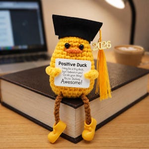 Könnte beinhalten: Eine gehäkelte gelbe Ente mit schwarzer Abschlusskappe und einem Schild mit der Aufschrift "Positive Duck". Die Ente hat braune Beine, gelbe Schuhe und eine goldene Quaste mit "2026". Die Ente sitzt auf einem geschlossenen Buch.
