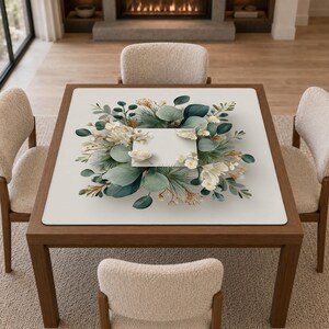 May include: A square table mat featuring a central botanical illustration of eucalyptus leaves, pine needles, and white flowers arranged in a circular wreath pattern around a blank white square space on a solid off-white background.