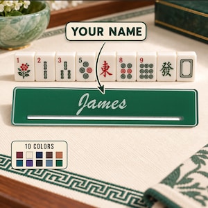 May include: A rectangular green Mahjong tile rack featuring the custom name James in white script font. The rack is displayed with a row of eight white Mahjong tiles with various red, green, and black symbols, plus a color chart showing ten options.