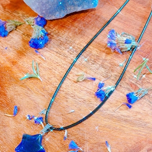 May include: A dark blue star pendant necklace with a black cord. The star is a deep blue color and is set against a wooden background scattered with blue flower petals and a large blue crystal.