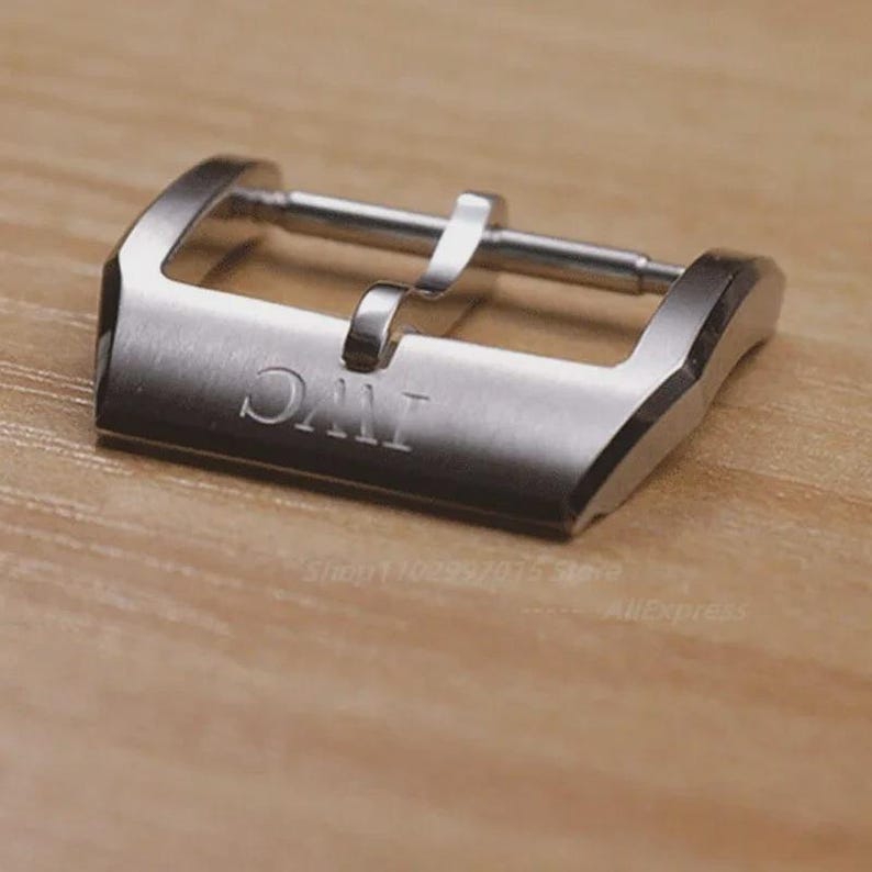 Puede incluir: Una hebilla de reloj plateada con las letras "IWC" grabadas. La hebilla tiene forma rectangular con un dise&ntilde;o curvo y un cierre de pasador. El fondo es una superficie de madera marr&oacute;n claro.